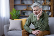 © Liubomir - Mature man with gray beard sitting on a couch, holding his ear in discomfort, grimacing from ear pain or tinnitus in a modern living room, appearing worried and tired