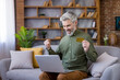 © Liubomir - Mature bearded man with gray hair celebrates online shopping success on his laptop at home, holding a gold credit card with a joyful, satisfied expression on the sofa