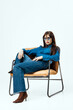 © wedmoments.stock - Young woman with long brown hair wearing blue shirt and sunglasses sits casually on a modern chair against a white background, showcasing a stylish fashion look