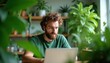 © miss irine - Man with beard works on laptop in green office. Computer screen glows, reflects light. Room filled with houseplants, creating nature vibe. Person types on keyboard at desk.