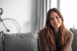 © luciano - Smiling young woman sitting on a sofa in a cozy living room, wearing a casual sweater and looking relaxed and approachable; natural light and warm tones create a comfortable, friendly atmosphere.