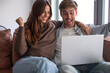 © luciano - A young couple sits on a sofa with arms around each other, excitedly celebrating while looking at a laptop screen in a bright, casual living room setting that conveys joy and connection.