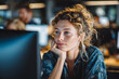© EricMiguel - Young female software developer with freckled skin and curly blonde bun, resting chin on hand while gazing at monitor in warm open - plan office