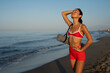 © sutulastock - Cheerful athletic woman in red sportswear smiling joyfully on a beach during sunrise, holding a yoga mat and enjoying a moment of happiness and freedom by the ocean