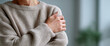 © perfectlab - Close-up of person wearing cozy beige knitted sweater holding their shoulder indoors with blurred background
