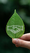 © Jan - Conceptual vertical image of a hand holding a green leaf with a handshake symbol etched on it, representing environmental partnership, eco-friendly business deals, sustainable cooperation, and natu...