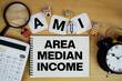 © may1985 - AMI Area Median Income text written on notepad with house model, alarm clock and magnifying glass on wooden background