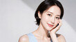 © YT - やわらかな斜光が差し込むグレー背景の前で頬に手を添えて微笑むショートヘアの若い日本人女性。淡いブルーのノースリーブと透明感のあるツヤ肌が印象的な、スキンケア・美容広告向けの清潔感あふれるポートレート素材
