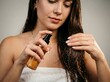 © Iwan - A young woman in a white towel applying a hair product from a spray bottle to her wet, dark hair after showering, focusing on hair care.