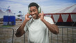 © Krakenimages.com - Man holds large overear headphones to his ears with both hands, smiling in front of red white striped circus tent and metal barrier at a street fair; joyful listening.