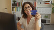 © Krakenimages.com - Woman holding blue creditcard with hand, smiling at monitor while seated at desk in building; confidence convenience.