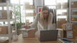 © Krakenimages.com - Woman leaning over laptop and signing shipping label on cardboard box amid stacked parcels and shelves in studio; small business focused.