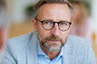 © lashkhidzetim - Man with glasses and beard looks directly at camera. He wears a light blue shirt and grey blazer. Background softly blurred with warm tones. His expression is calm, thoughtful, and professional