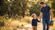 © Yulia - Dad carrying child with a colorful backpack on a sunny forest trail, both smiling and enjoying a nature walk, surrounded by lush greenery and warm sunlight