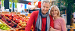 © Prostock-studio - A couple stands together at a market with baskets of fruit around them. They are smiling while enjoying their time shopping for fresh produce. Colorful fruits are displayed in the background.