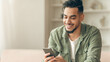 © Prostock-studio - A young man sits on a couch at home, looking at his smartphone with a smile. He is dressed casually and appears to enjoy his time on the device. The room has soft lighting and simple decor.