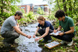 © AiHRG Design - Students collect water samples from a stream for research. They wear casual clothes and work near a university building. The scene shows hands-on learning in nature.