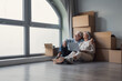© Daniel - Elderly couple sitting on the floor surrounded by moving boxes, using a laptop, and looking out a large window, contemplating life in their new apartment or home