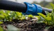© Varos - A close-up shot of a black irrigation pipe with a blue coupling connector dripping a clear water droplet onto the dark, moist soil of a vegetable garden.