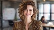 © Muntazim - A smiling young Caucasian woman with curly hair, wearing a floral blouse, radiates confidence in a bright office setting.
