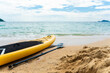 © Kawee - A yellow paddleboard rests on the sandy beach beside the calm sea.