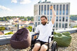 © sofiko14 - A man with a beard and long hair in a wheelchair raises his fist in triumph on a rooftop terrace with a city view
