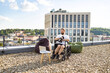 © sofiko14 - A man in a wheelchair is exercising with dumbbells on a rooftop terrace while watching a laptop