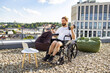 © sofiko14 - A man with a beard in a wheelchair is exercising with dumbbells on a rooftop terrace with a city view