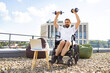 © sofiko14 - A man with a beard sits in a wheelchair on a rooftop, lifting dumbbells while looking at a laptop screen, with beanbags and city buildings in the background
