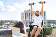 © sofiko14 - A man with a beard and long hair in a wheelchair is exercising with dumbbells on a rooftop terrace with a laptop and beanbag chair nearby