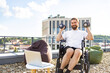 © sofiko14 - A man with a beard in a wheelchair is exercising with dumbbells on a rooftop terrace with a laptop and beanbag chair nearby