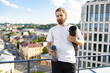© sofiko14 - A bearded man in a white t-shirt stands on a rooftop holding a yoga mat and a water bottle with a city view behind him