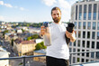 © sofiko14 - A man with a beard and bun hairstyle holds a water bottle and a rolled-up yoga mat on a rooftop overlooking a city skyline