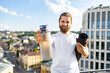 © sofiko14 - A smiling man with a beard and long hair holds a water bottle and a rolled-up yoga mat on a rooftop with a city view