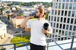 © sofiko14 - A bearded man with his hair in a bun drinks from a large water bottle while holding a rolled-up yoga mat on a rooftop overlooking a city
