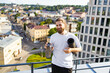 © sofiko14 - A smiling man with a beard and long hair holds a yoga mat and a water bottle on a rooftop overlooking a city