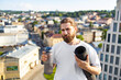 © sofiko14 - A man with a beard and long hair holds a water bottle and a rolled-up yoga mat while standing on a rooftop overlooking a city