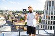© sofiko14 - A man with a beard and headphones holds a smartphone on a gimbal and a water bottle while filming himself on a city rooftop