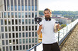 © sofiko14 - A man with a beard and headphones holds a water bottle and a smartphone on a selfie stick, filming himself on a rooftop with a city skyline in the background