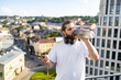 © sofiko14 - A man with headphones and a beard drinks from a water bottle while holding a selfie stick with a smartphone attached, overlooking a city skyline