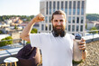 © sofiko14 - A smiling man with a beard and headphones around his neck flexes his bicep while holding a water bottle on a rooftop with a city view