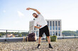 © sofiko14 - A man with a beard and tied-back hair performs a side stretch on a gravel rooftop with beanbag chairs and a modern building in the background