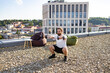 © sofiko14 - A man with a beard and tied-back hair performs dumbbell squats on a gravel-covered rooftop with a city view and modern building in the background