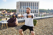 © sofiko14 - A smiling man with a beard and bun is performing bicep curls with dumbbells on a rooftop terrace with a city view
