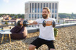 © sofiko14 - A smiling man with a beard and long hair is performing bicep curls with dumbbells on a rooftop terrace with a city view