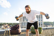 © sofiko14 - A man with a beard and bun hairstyle is performing a dumbbell workout on a rooftop terrace with a city view in the background