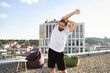 © sofiko14 - A man with a beard and bun hairstyle performs a shoulder stretch on a rooftop terrace with a city view and modern building in the background