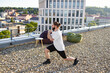 © sofiko14 - A man with a beard and bun hairstyle performs a lunge exercise on a gravel rooftop with a city view and beanbag chairs