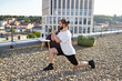 © sofiko14 - A man with a beard and bun performs a lunge exercise on a gravel-covered rooftop with a city skyline in the background