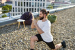 © sofiko14 - A bearded man with his hair in a bun performs a lunge exercise on a gravel-covered rooftop with city buildings in the background